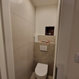 Tom-Fliz - Nowoczesna toaleta w trakcie wykańczania: beżowe płytki, podwieszana muszla, widoczne rury i narzędzia. Oświetlenie sufitowe.