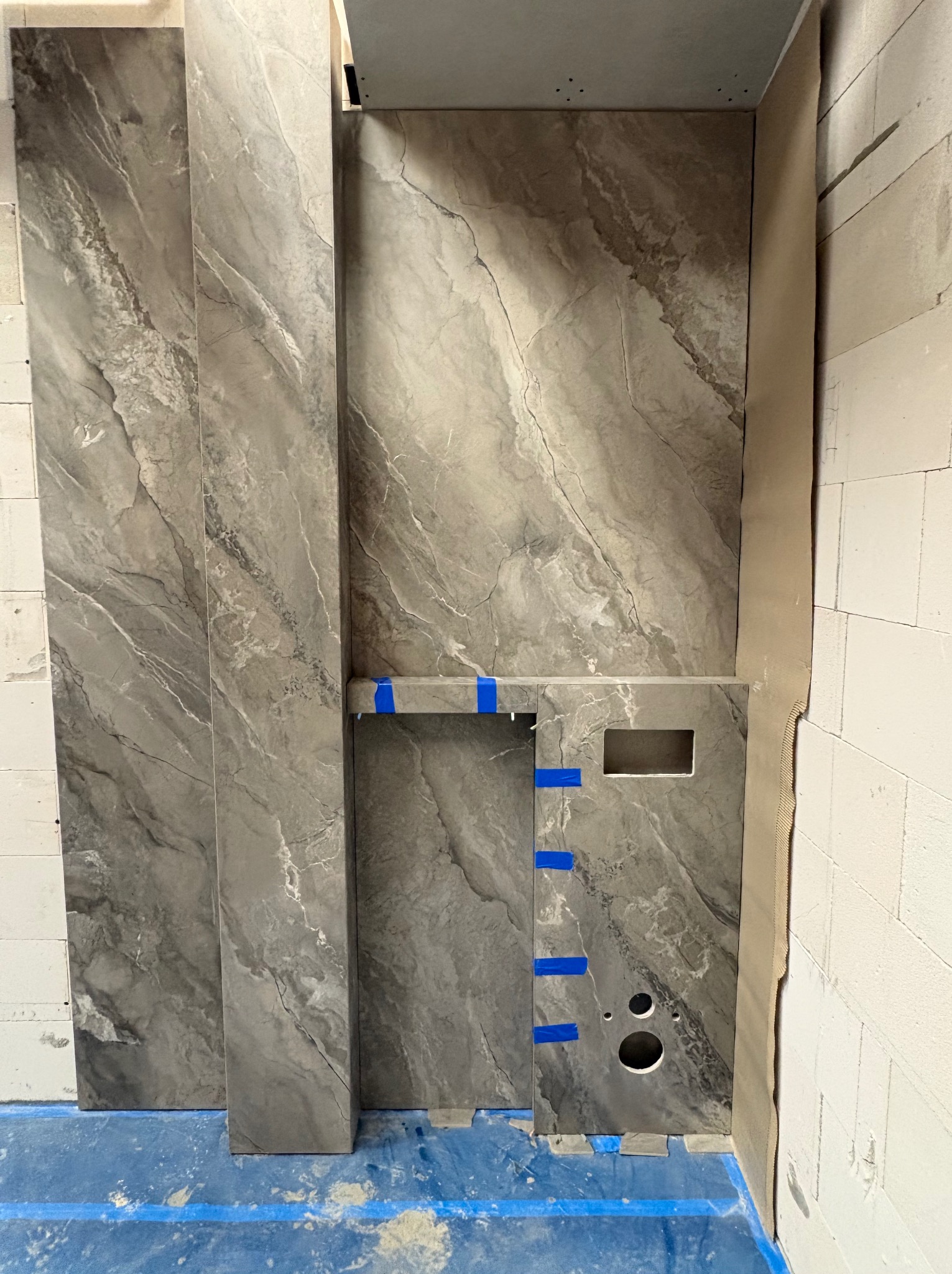 Montaż płytek imitujących marmur w łazience, widoczne otwory na instalacje i niebieska taśma zabezpieczająca. Prace glazurnicze w toku, surowy stan.