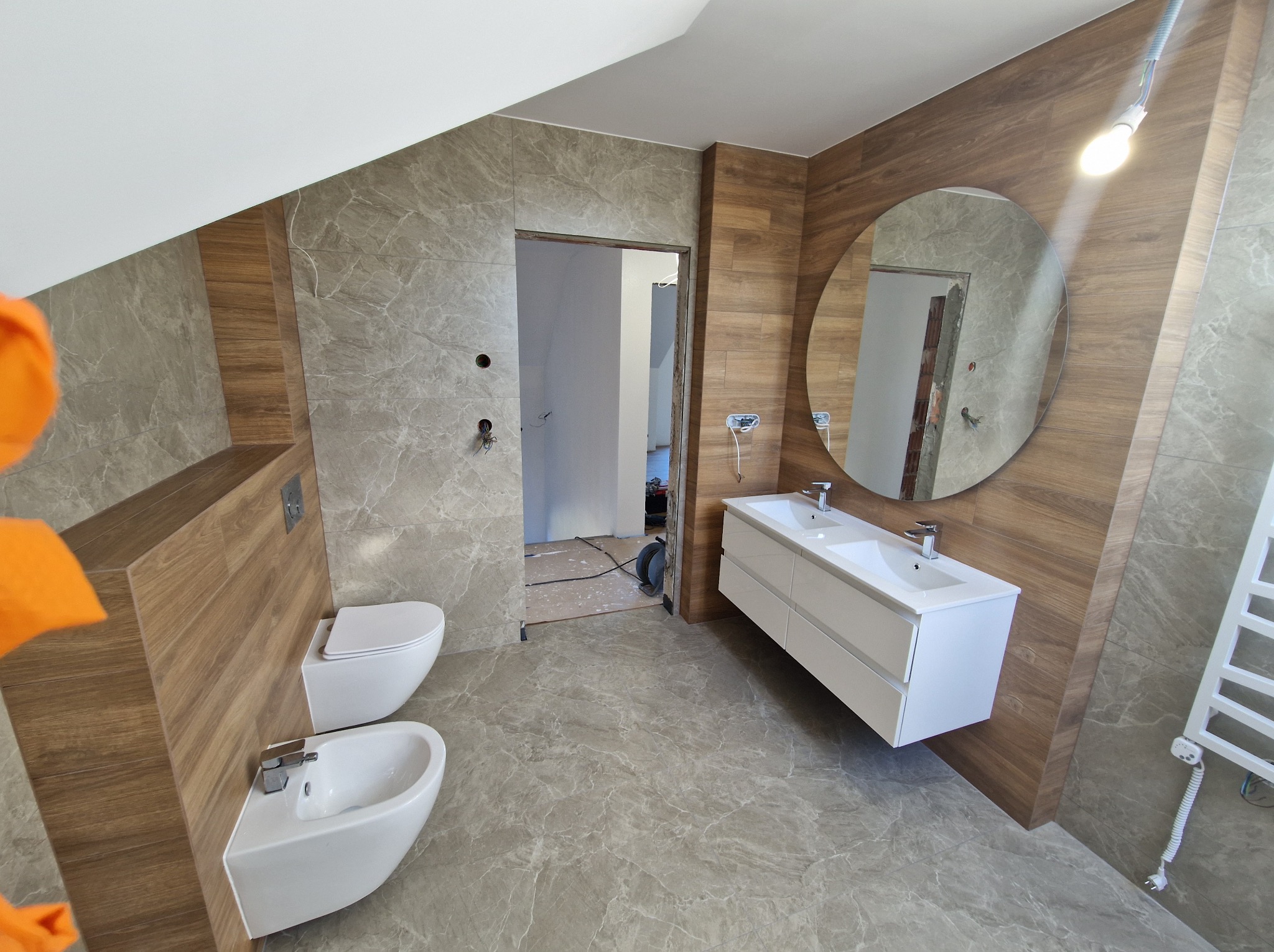 Nowoczesna łazienka w odcieniach szarości i drewna, z owalnym lustrem, podwieszaną toaletą i bidetem. Widoczne płytki imitujące marmur i drewno.