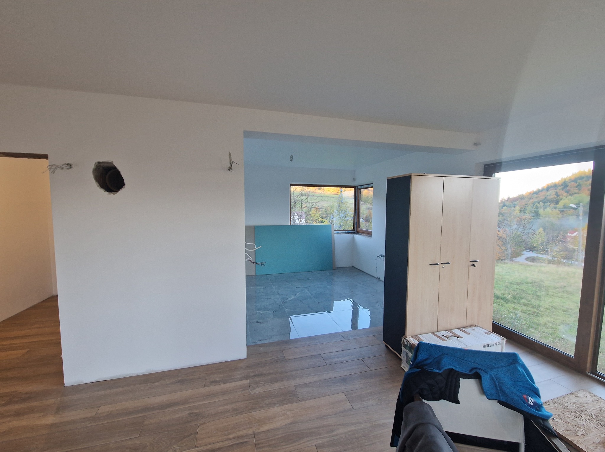 Wnętrze w trakcie prac wykończeniowych: drewnopodobna podłoga, gładkie ściany z otworem na instalację, w tle płytki i okno z widokiem na zieleń.