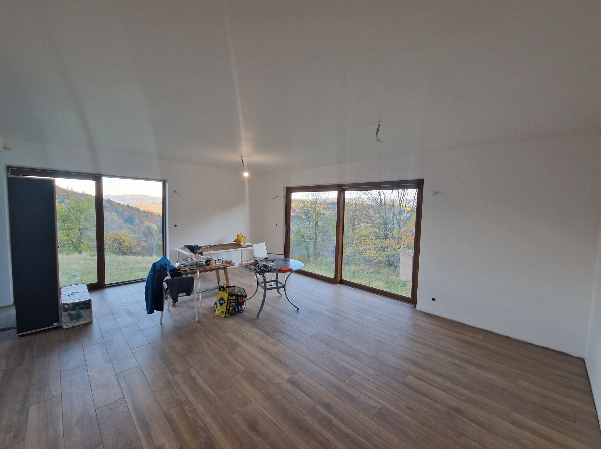 Jasne wnętrze z drewnopodobną podłogą i dużymi oknami z widokiem na zalesione wzgórza. Widoczne narzędzia i materiały sugerują etap prac wykończeniowych.