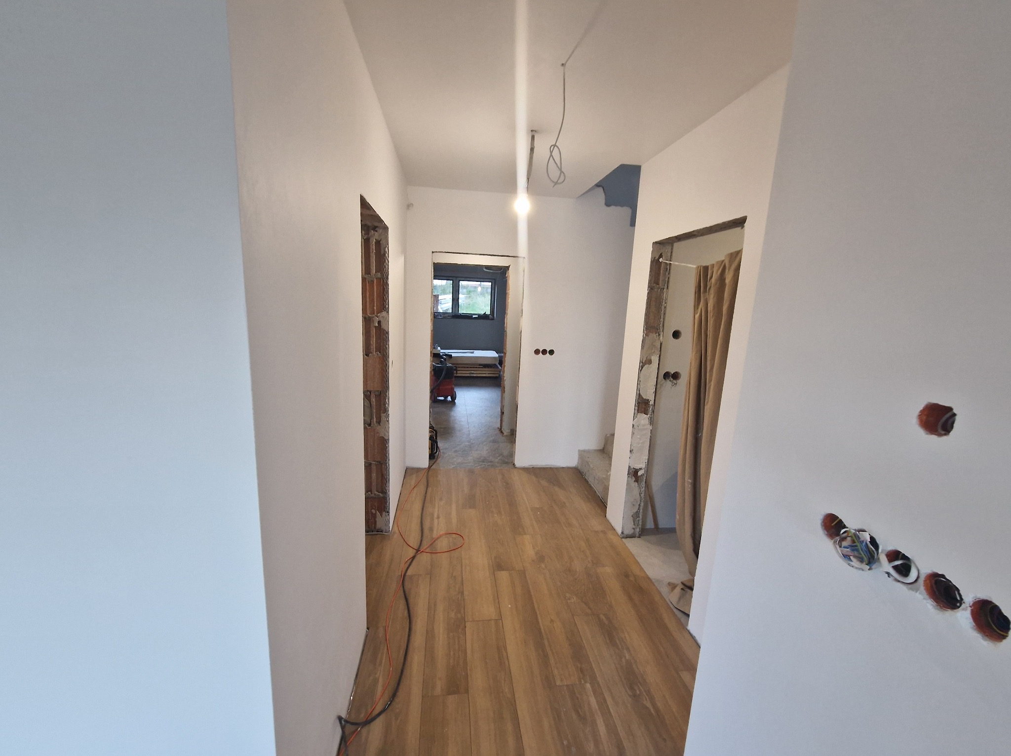 Korytarz w trakcie prac wykończeniowych: świeżo pomalowane ściany na biało, drewnopodobna podłoga, widoczne instalacje elektryczne i otwory drzwiowe bez ościeżnic.