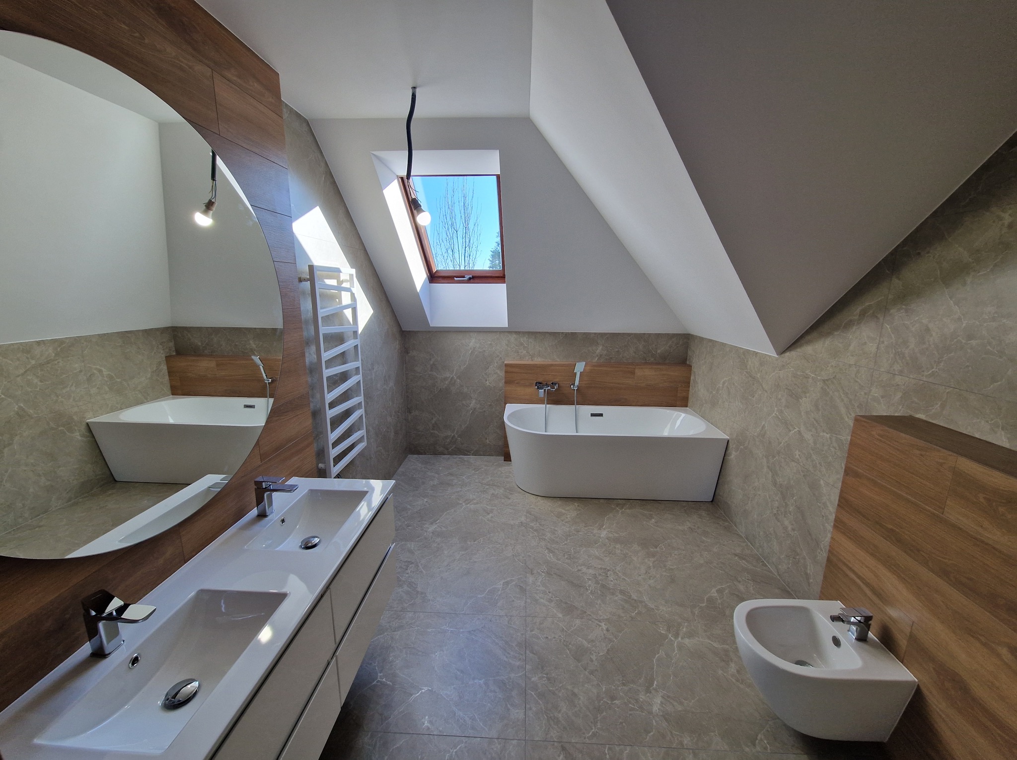 Nowoczesna łazienka na poddaszu z wanną wolnostojącą, umywalką nablatową i bidetem. Uwagę zwraca duże lustro o zaokrąglonym kształcie i okno dachowe.