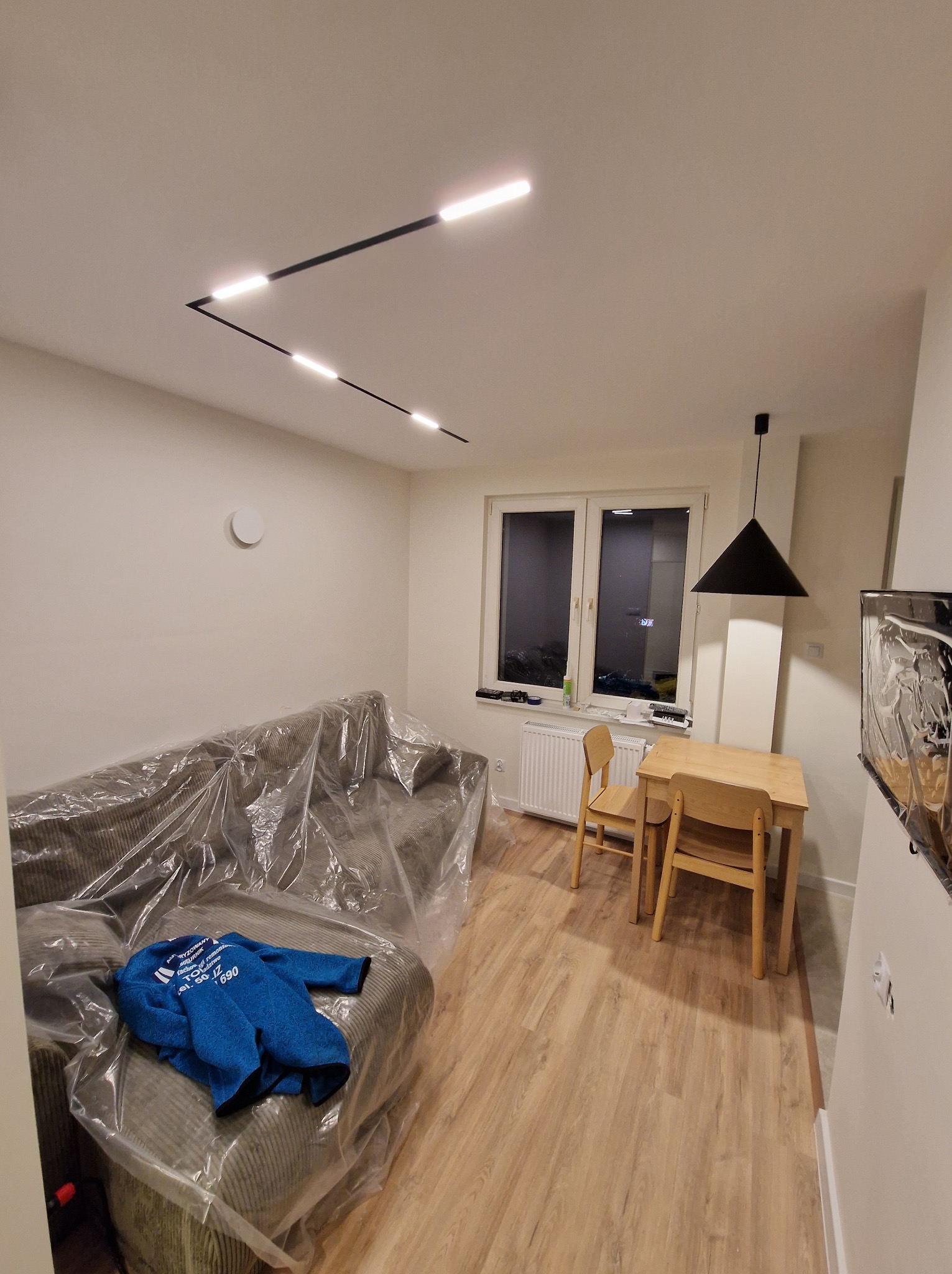 Jasne wnętrze pokoju po odświeżeniu: parkiet, stół z krzesłami, sofa przykryta folią ochronną, nowoczesne oświetlenie sufitowe. Widok na okno.