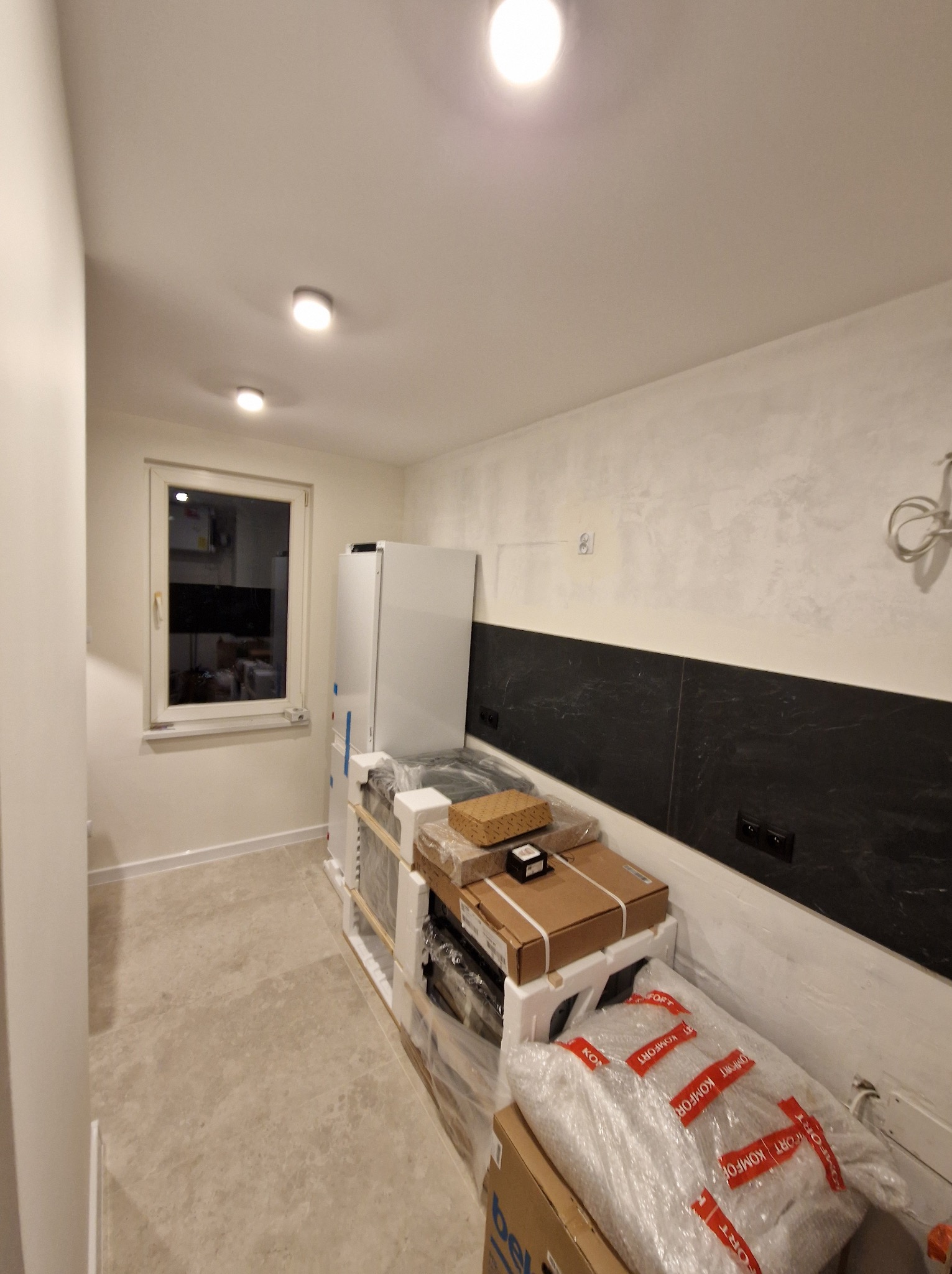 Nowa, jeszcze zapakowana kuchnia w trakcie montażu. Widoczne sprzęty AGD w folii ochronnej, czarne płytki na ścianie i jasne ściany. Przygotowanie do wykończenia wnętrza.