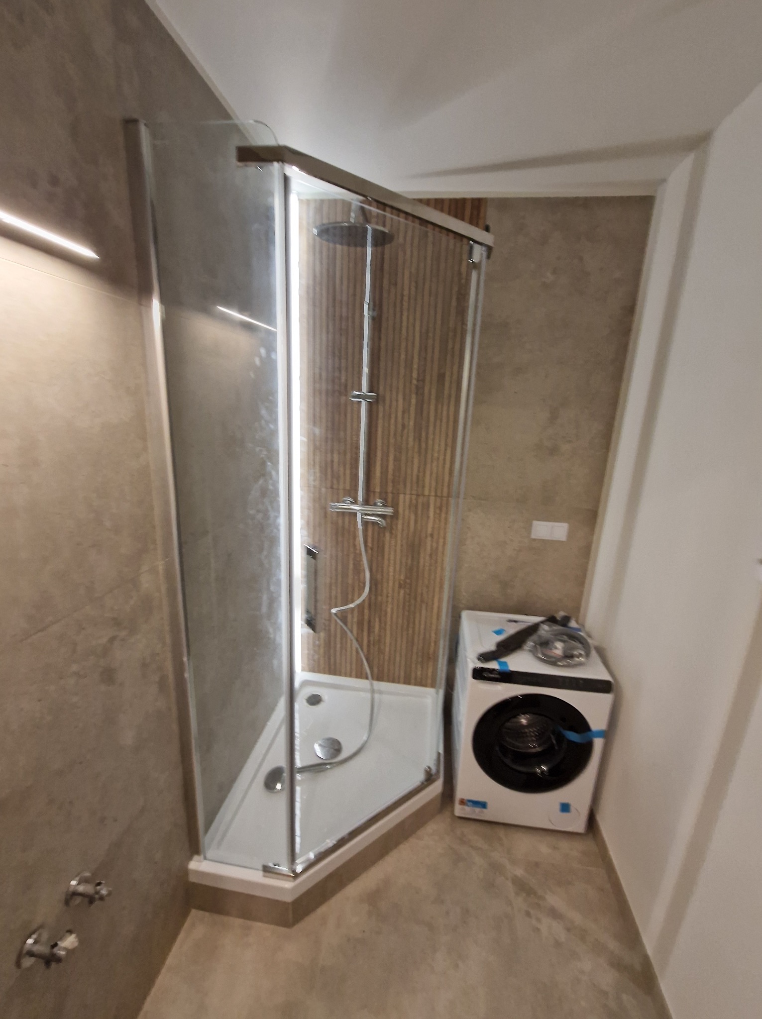 Nowoczesna kabina prysznicowa z deszczownicą w wykończonej łazience, obok pralka. Ściany wyłożone płytkami imitującymi drewno. Minimalistyczny design.