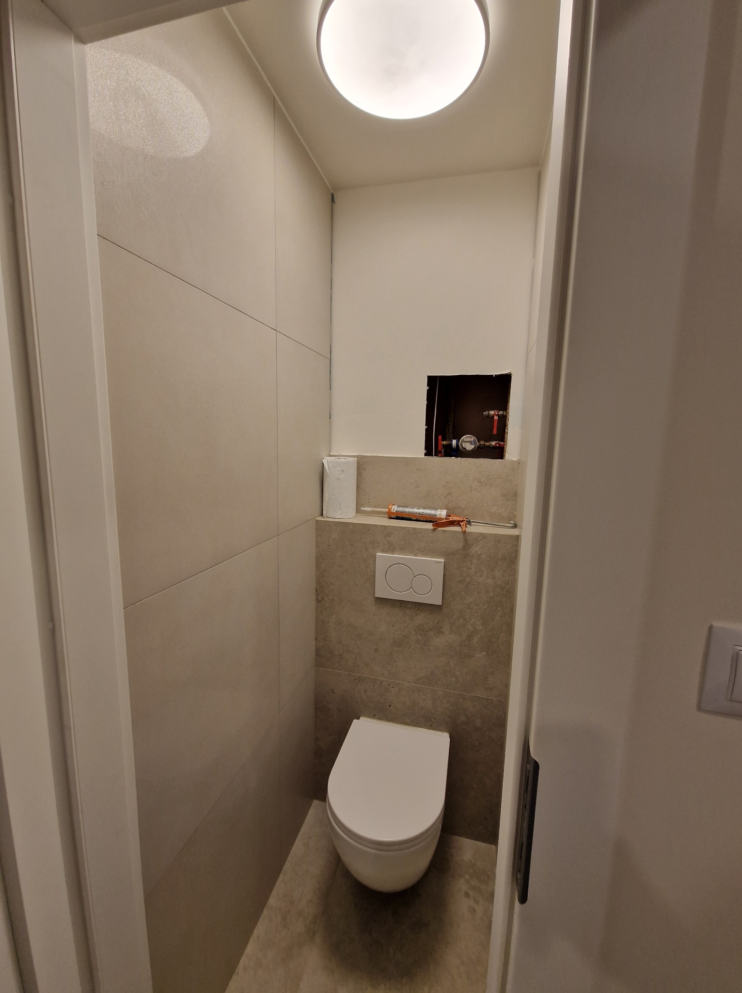 Nowoczesna toaleta w trakcie wykańczania: beżowe płytki, podwieszana muszla, widoczne rury i narzędzia. Oświetlenie sufitowe.