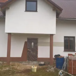 Biała elewacja domu jednorodzinnego z brązowym dachem i stolarką okienną, widoczne materiały budowlane i zabezpieczone folią drzwi wejściowe, wskazujące na trwające prace wykończeniowe.