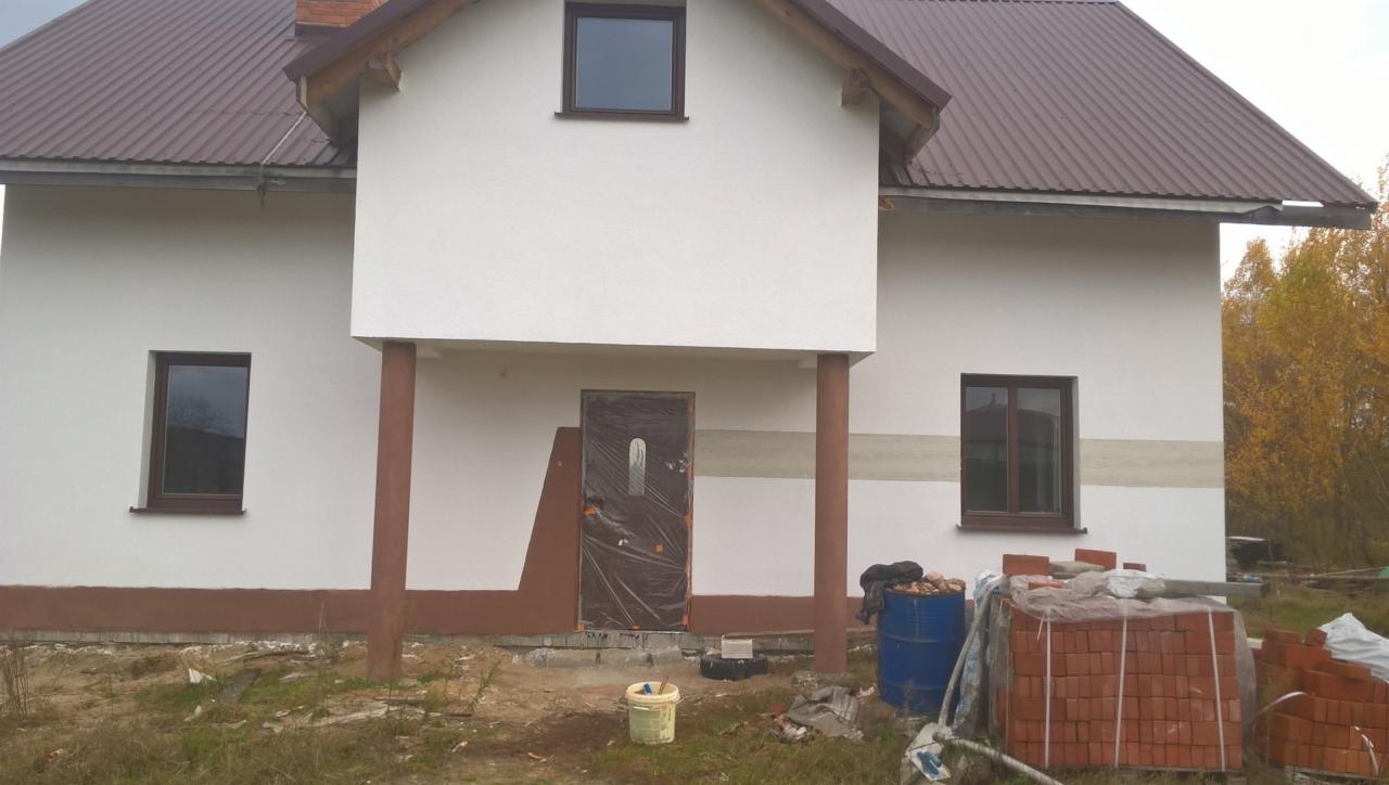 Biała elewacja domu jednorodzinnego z brązowym dachem i stolarką okienną, widoczne materiały budowlane i zabezpieczone folią drzwi wejściowe, wskazujące na trwające prace wykończeniowe.