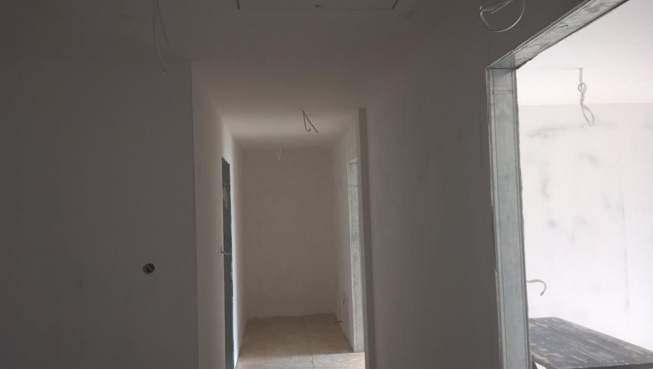 Wnętrze w trakcie wykańczania: ściany otynkowane na szaro, widoczne wystające przewody elektryczne i surowa ościeżnica drzwiowa z metalu.