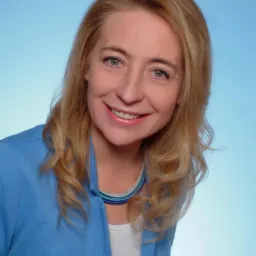 Portret uśmiechniętej kobiety w niebieskiej marynarce i naszyjniku z niebieskich koralików na jasnoniebieskim tle.