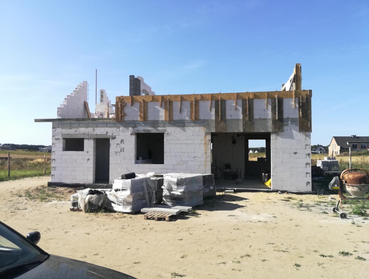 Niedokończony budynek jednorodzinny z białych bloczków, widoczne drewniane elementy konstrukcyjne dachu, na pierwszym planie materiały budowlane i betoniarka.