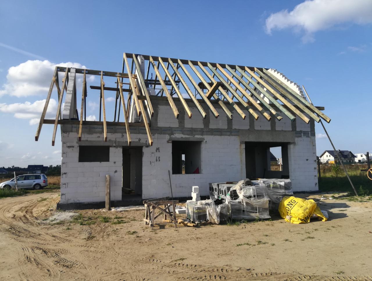 Budowa domu w stanie surowym otwartym: ściany z białego bloczka, drewniana konstrukcja dachu, materiały budowlane na placu budowy, widok na okoliczne domy w tle.