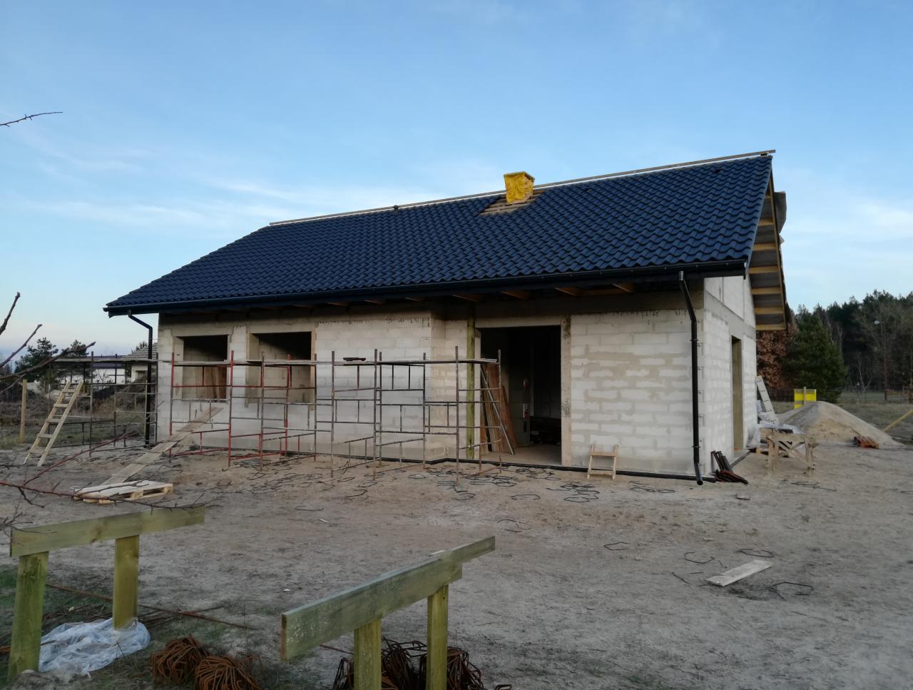 Budowa domu jednorodzinnego w stanie surowym otwartym: ściany z bloczków, ciemna dachówka, widoczne rusztowania i przygotowany teren.