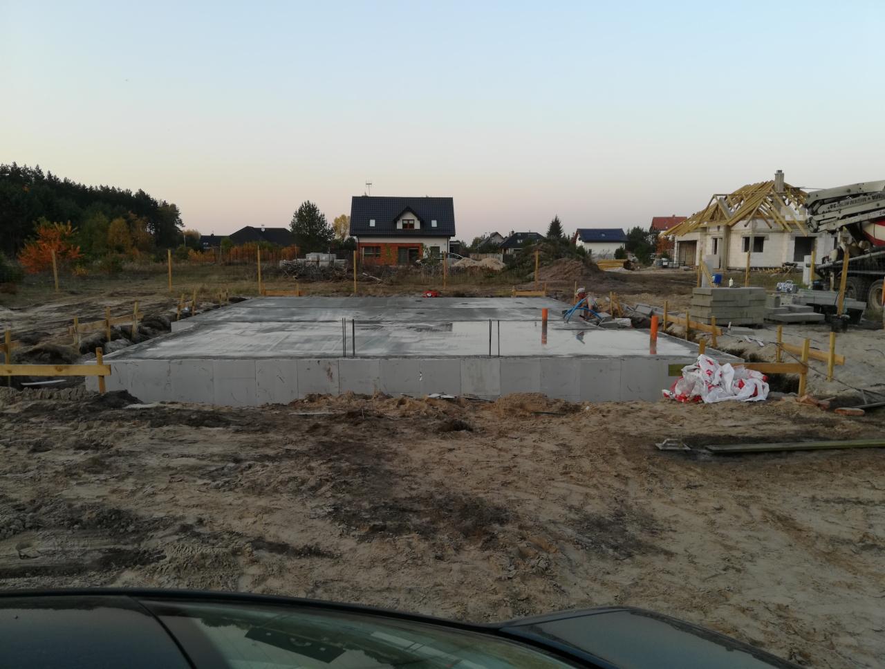 Wylana płyta fundamentowa z wystającymi prętami zbrojeniowymi, otoczona drewnianym szalunkiem, w tle dom w trakcie budowy z widoczną konstrukcją dachu i pompą do betonu.