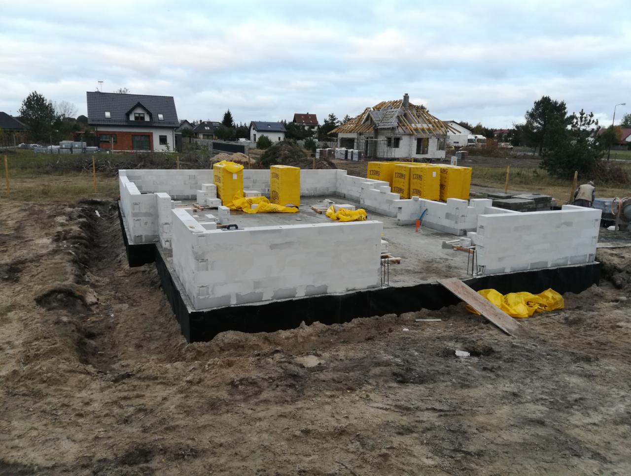 Fundamenty domu z bloczków Ytong, zabezpieczone izolacją, na działce w trakcie budowy z widocznym nowym domem w tle.