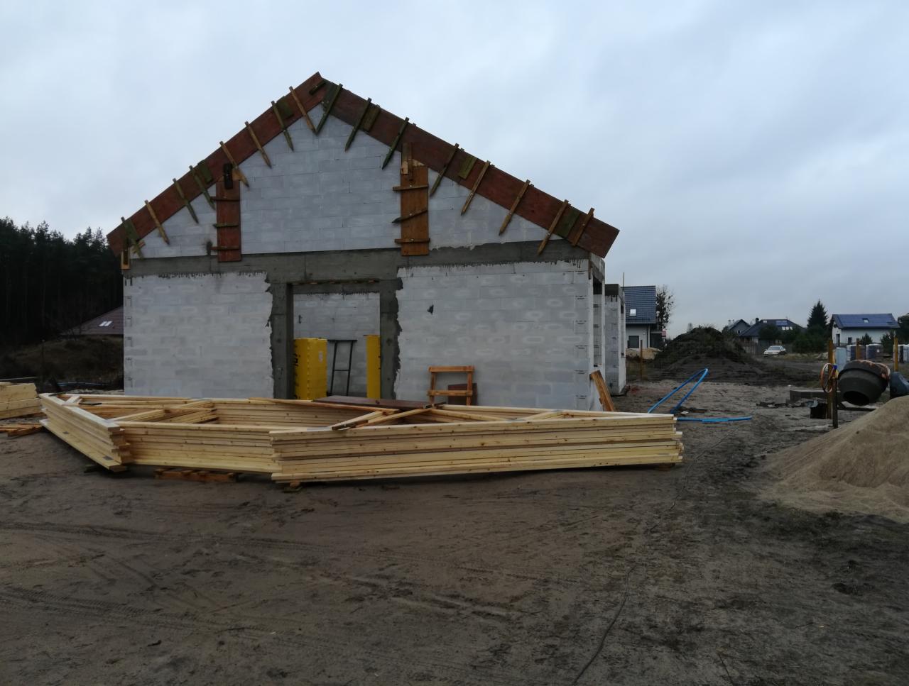 Surowy stan domu z betonu komórkowego z drewnianą konstrukcją dachu w trakcie budowy, widoczne przygotowane elementy więźby dachowej, na placu budowy stoi betoniarka.