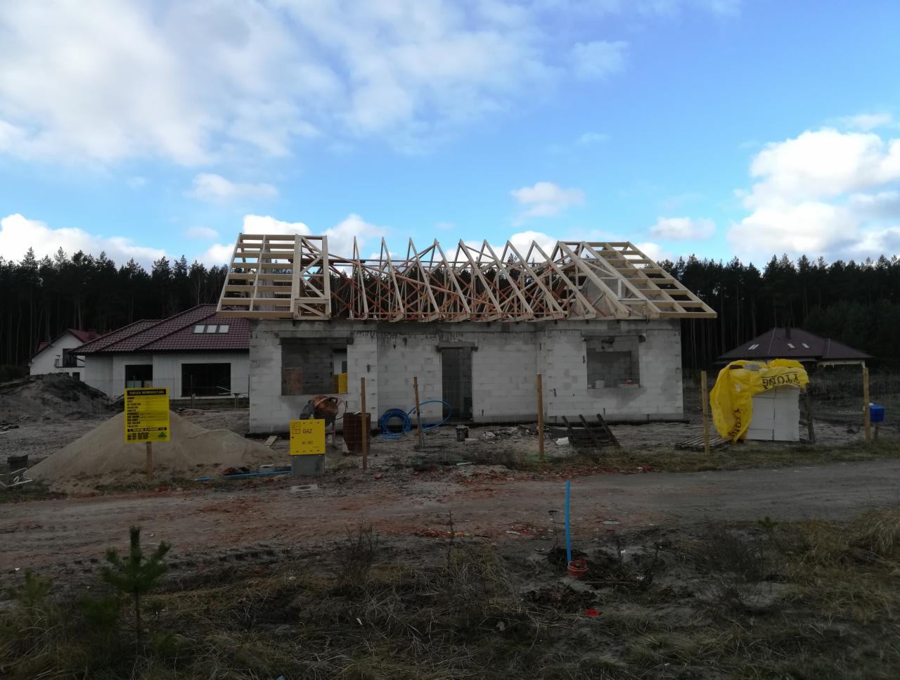 Surowy stan domu jednorodzinnego z pustaków z drewnianą konstrukcją dachu w trakcie budowy, widoczne oznakowanie przyłącza gazu, w tle las