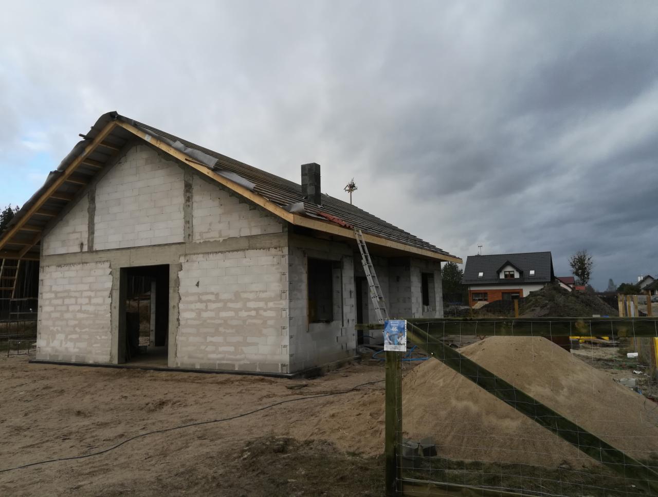 Wznoszenie ścian domu jednorodzinnego z bloczków betonu komórkowego, widoczna konstrukcja dachu i drabina oparta o budynek na tle pochmurnego nieba.