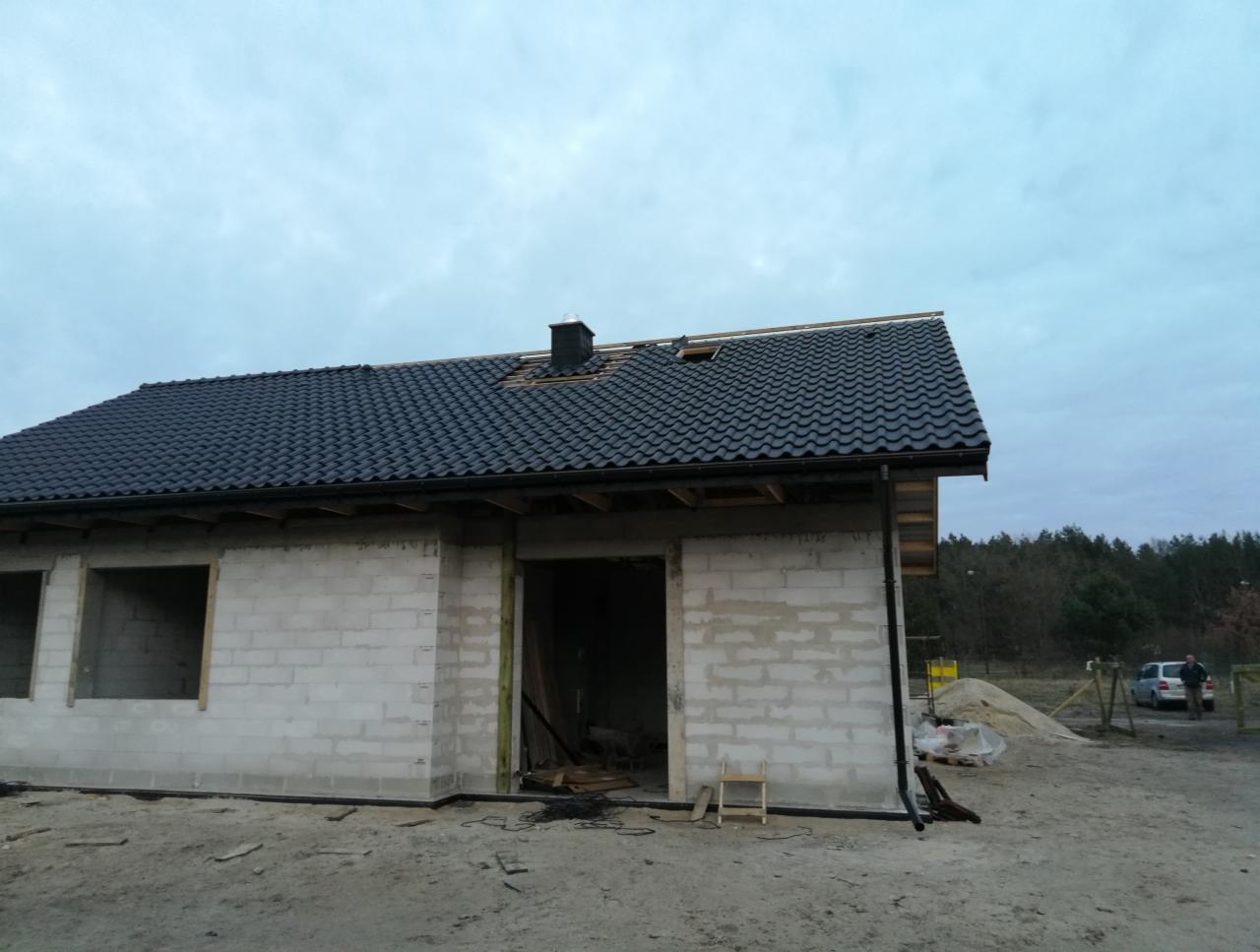 Surowy stan domu jednorodzinnego z czarną dachówką, widoczne bloczki betonowe, otwory okienne i drzwiowe, teren budowy z piaskiem i drewnianymi elementami szalunkowymi.