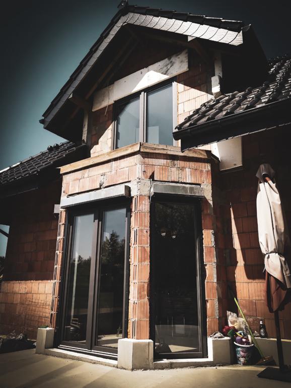 Dom w trakcie budowy z zamontowanymi oknami i drzwiami z profili Salamander, widoczne elementy konstrukcyjne z czerwonej cegły, betonowe podstawy pod oknami oraz rozłożony parasol w tle.