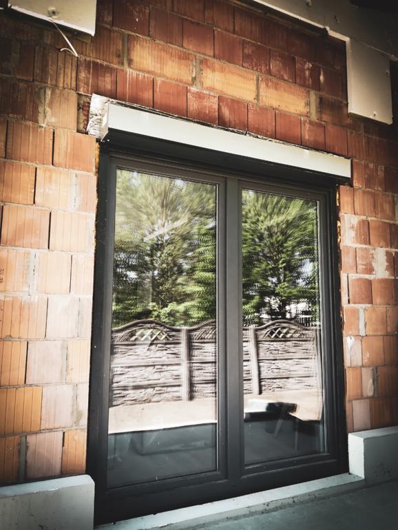 Zamontowane dwuskrzydłowe okno PCV w surowej ścianie z cegły ceramicznej, widoczna kaseta rolety zewnętrznej nad oknem, odbicie ogrodu w szybie.