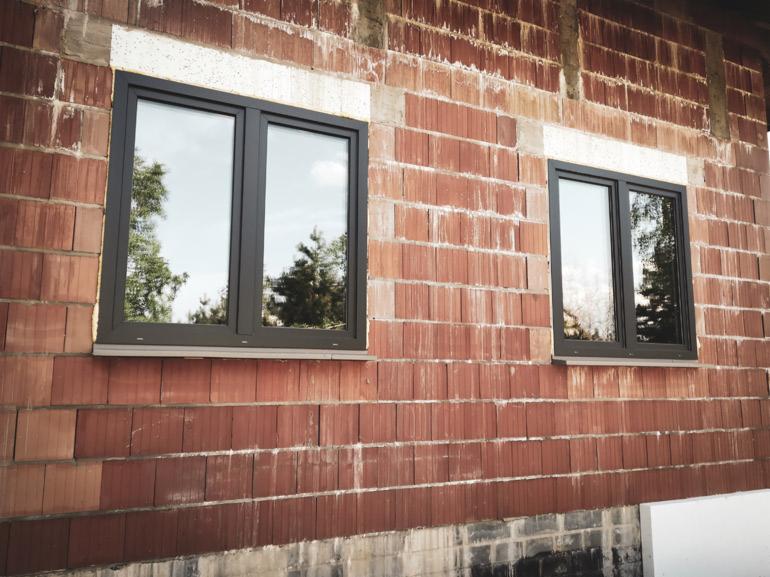 Dwa okna PCV z ciemnymi ramami zamontowane w surowej, czerwonej cegle budynku w Częstochowie, z widocznym odbiciem drzew w szybach.