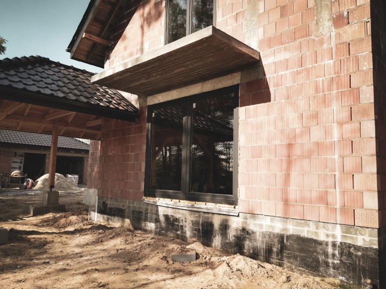 Montaż okna PCV z czarnym profilem w surowym budynku z czerwonej cegły, widoczna betonowa nadproże i przygotowany teren wokół.