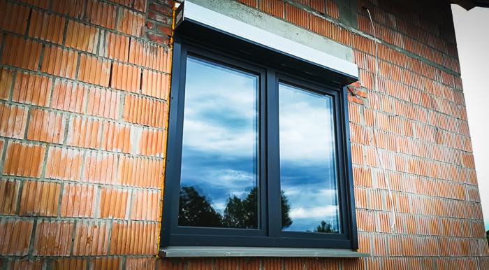 Nowe okno PCV z roletą zewnętrzną zamontowane w surowej ścianie z czerwonej cegły, widok na odbicie nieba i drzew w szybach, profil Veka Softline.