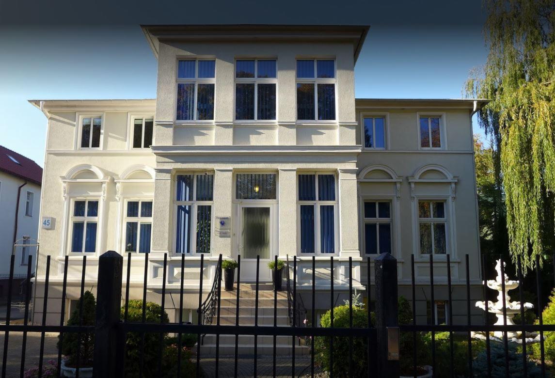 Elewacja trzypiętrowego budynku biurowego z licznymi oknami, widoczna zza czarnego, metalowego ogrodzenia.