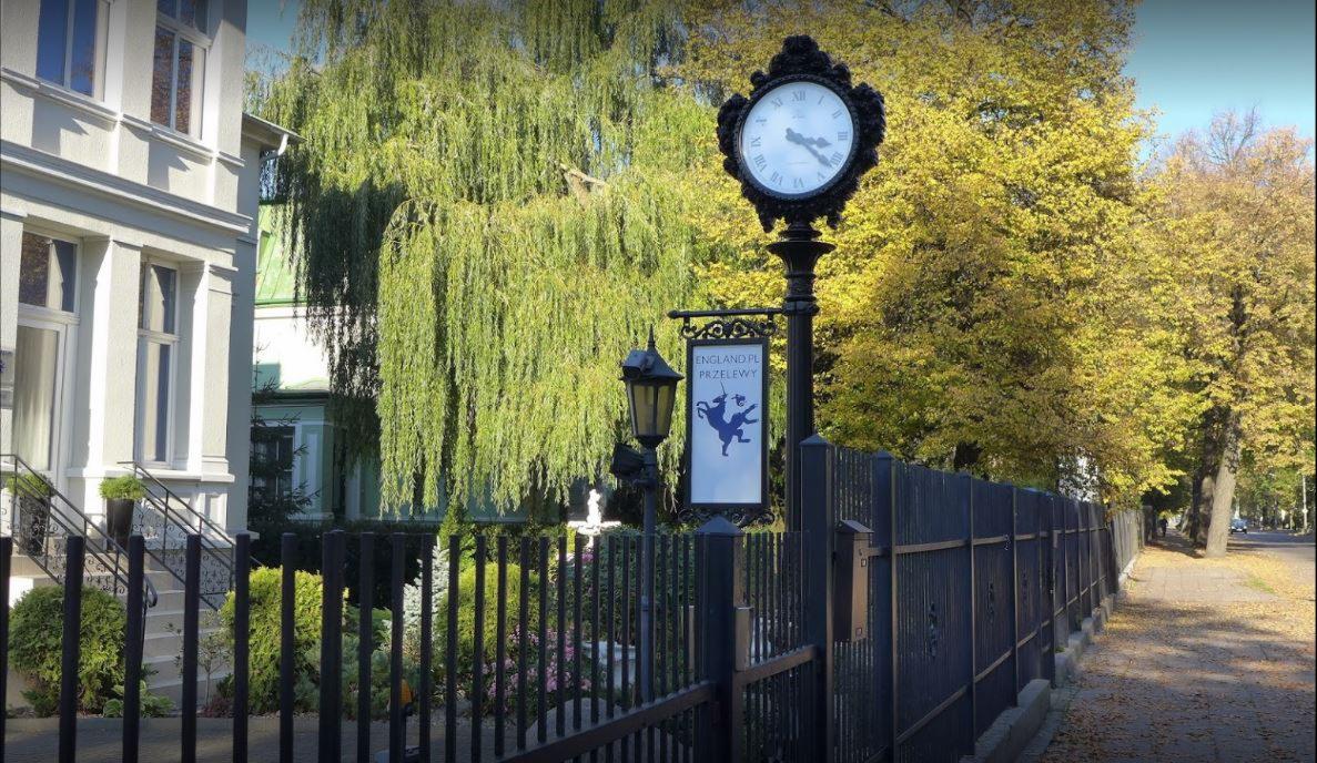 Elegancka siedziba firmy z zegarem na słupie, tablicą reklamową 'England.pl Przelewy' z motywem rycerza na koniu, widoczna część budynku z zielonymi drzewami w tle.