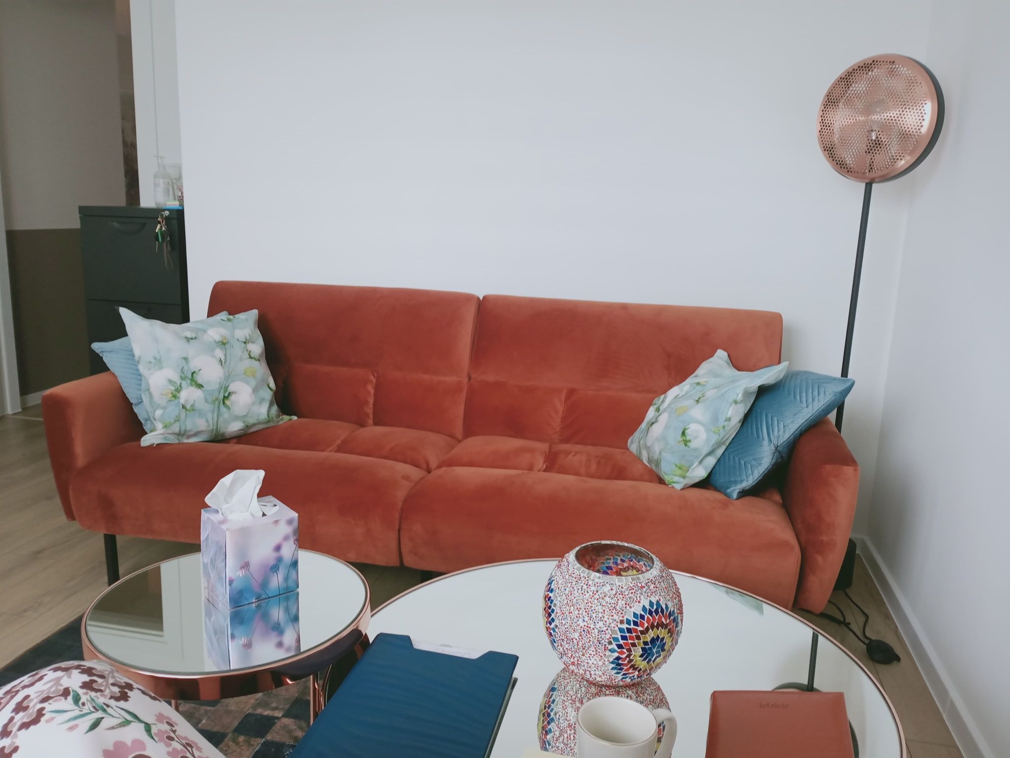 Pomarańczowa sofa z poduszkami w gabinecie, stoliki kawowe z dekoracjami, lampa podłogowa w tle, minimalistyczny wystrój.