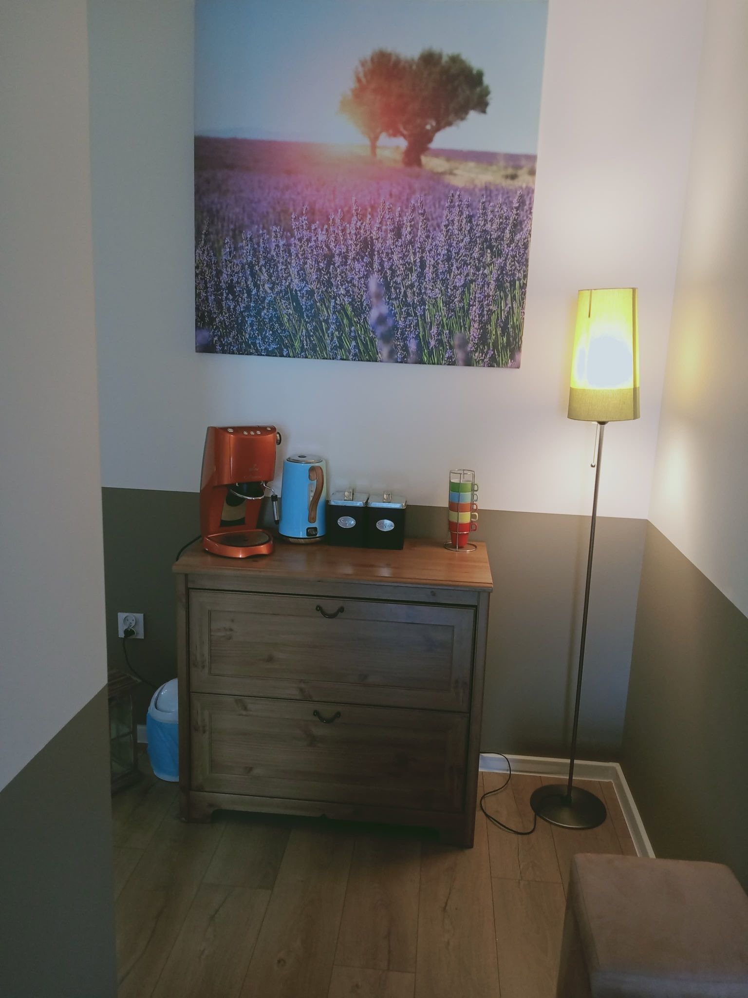Gabinet psychoterapeutyczny: drewniana komoda z ekspresem do kawy, czajnikiem i pojemnikiem na herbatę pod obrazem lawendy, obok lampa podłogowa i stos kolorowych kubków.