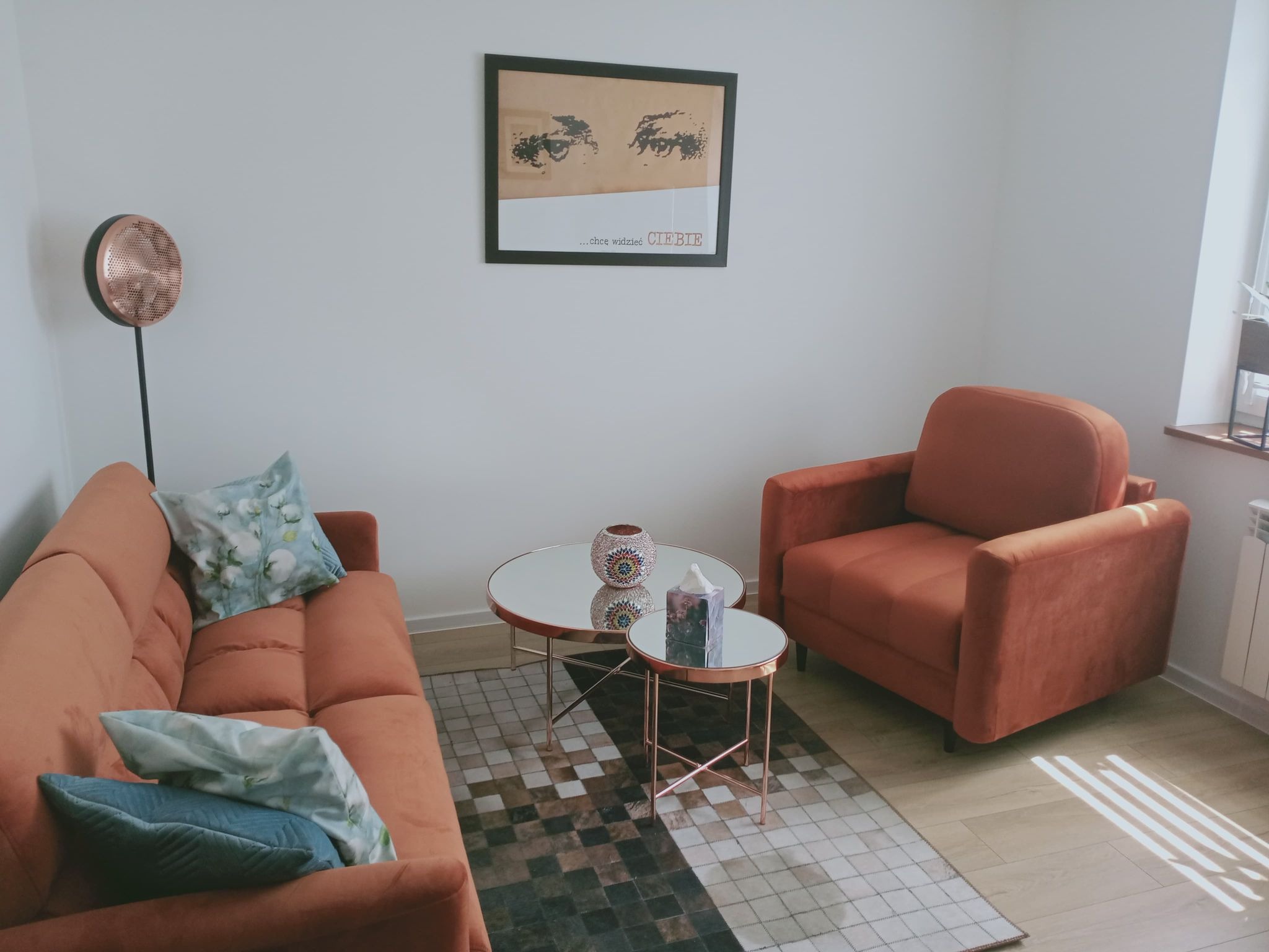 Pomarańczowy fotel i sofa z poduszkami w gabinecie z dwoma stolikami kawowymi z lustrzanymi blatami i obrazem na ścianie.