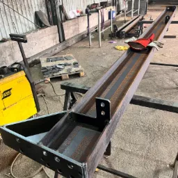 Metalowa konstrukcja z profili, spawana, z widocznym żółtym spawarkiem ESAB i rękawicami roboczymi w tle warsztatu. Detal z bliska, surowy stan.