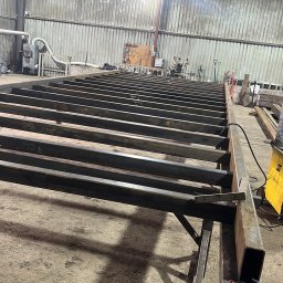 Piotr Trębicki - Konstrukcja stalowa w hali produkcyjnej: spawane profile tworzą szkielet, z narzędziami obok. Widoczne detale montażu i surowe oświetlenie.