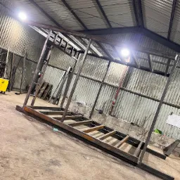 Konstrukcja stalowa hali w trakcie budowy, widoczna spawarka i fragment rysunku technicznego, surowy stan, oświetlenie przemysłowe.