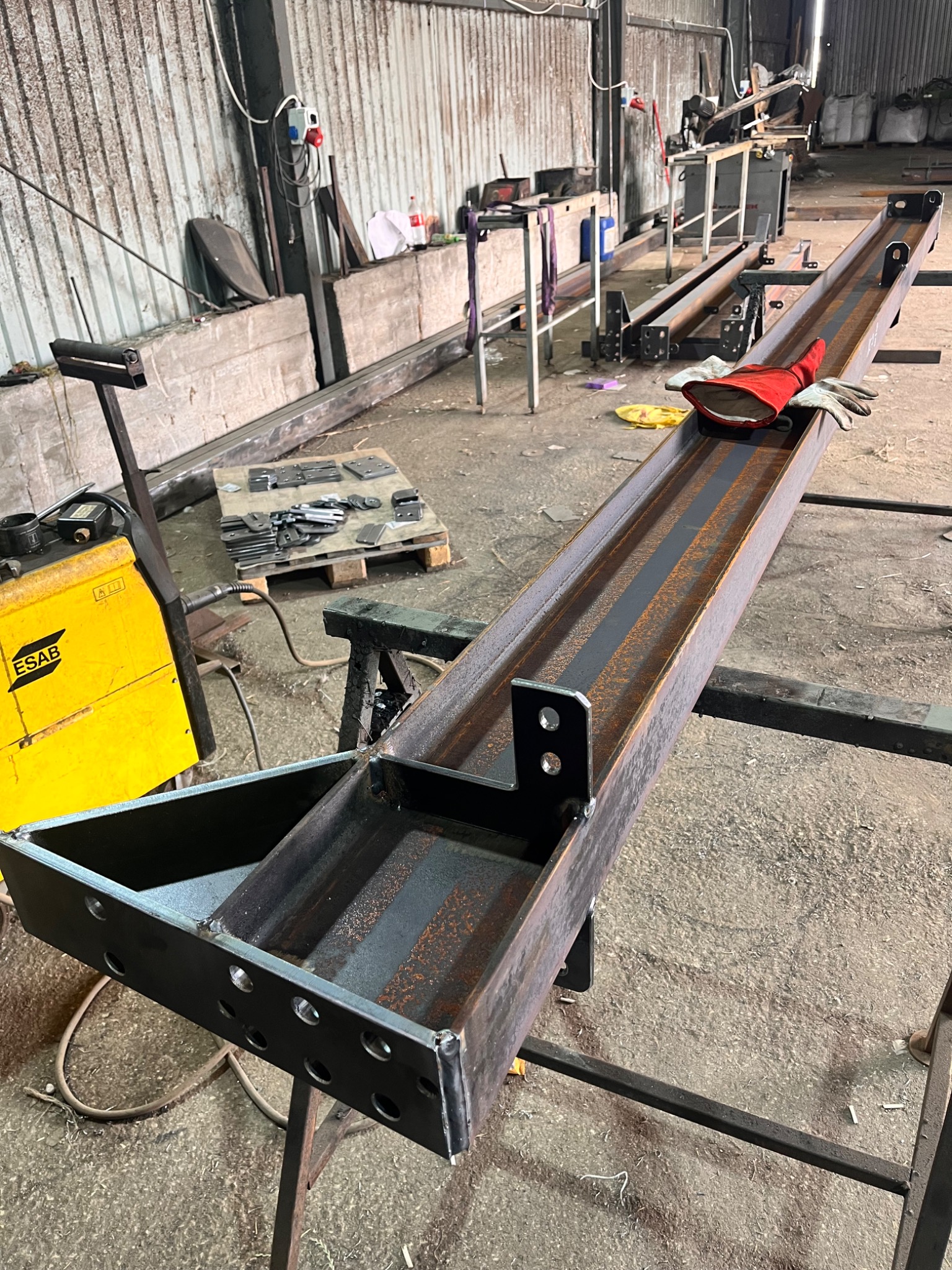 Metalowa konstrukcja z profili, spawana, z widocznym żółtym spawarkiem ESAB i rękawicami roboczymi w tle warsztatu. Detal z bliska, surowy stan.