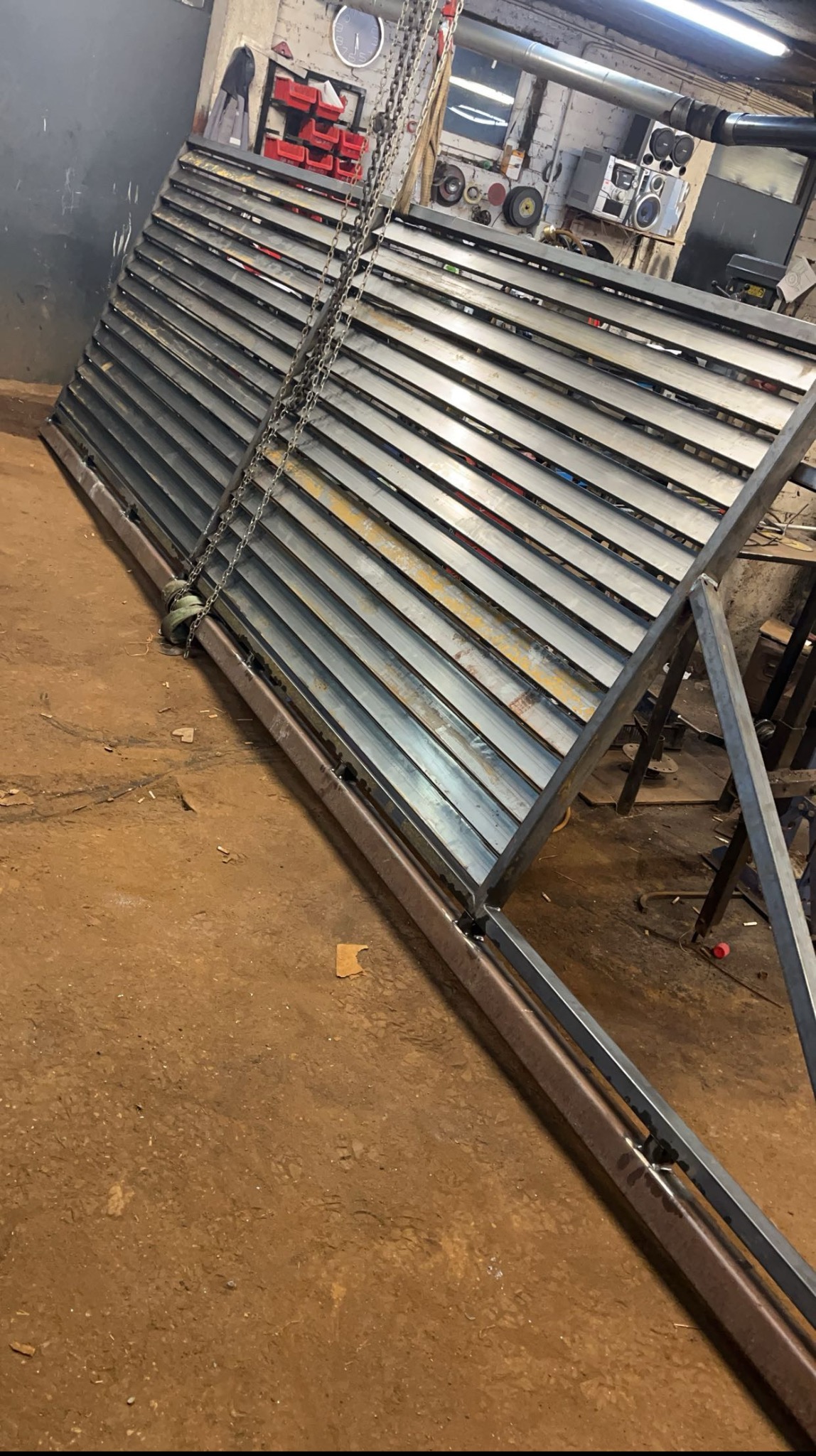 Konstrukcja metalowa z poziomymi lamelami, podwieszona na łańcuchach w warsztacie. Widoczne ślady spawania i obróbki, surowe wykończenie.