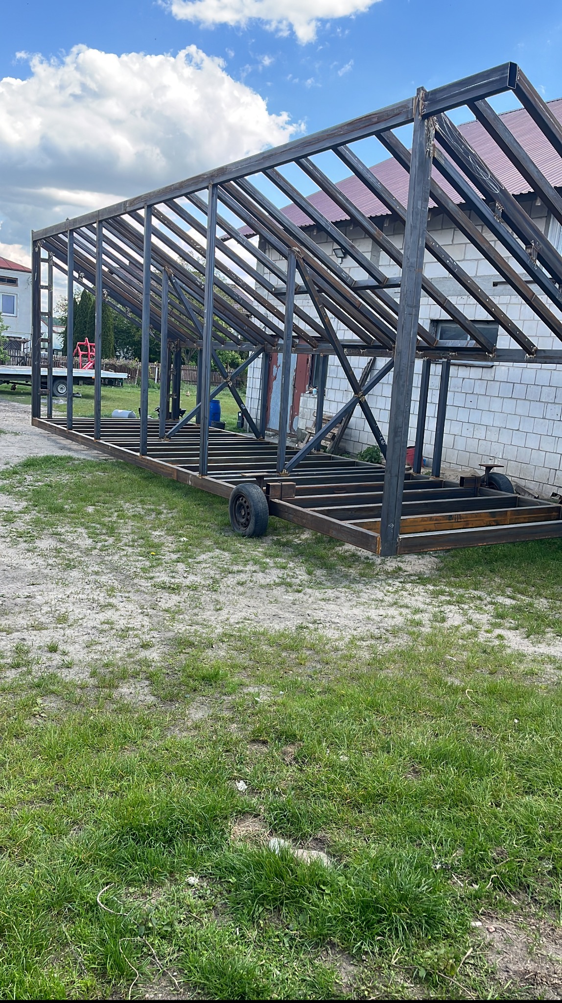 Konstrukcja stalowa hali w trakcie budowy, widoczne elementy szkieletu i podstawa na kołach, tło to budynek z białej cegły i fragment zielonego trawnika.