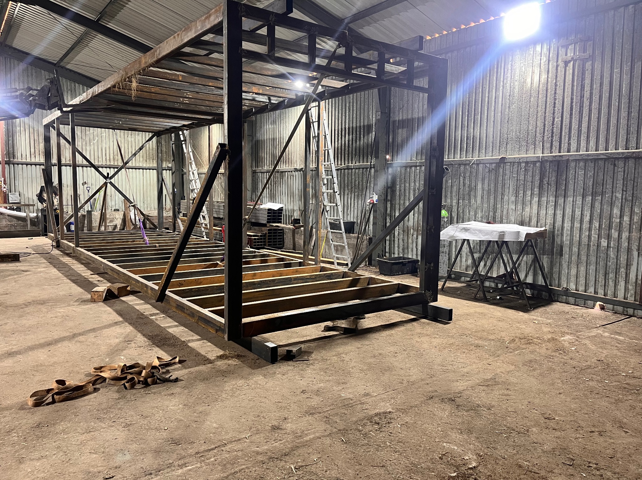 Konstrukcja stalowa hali w trakcie budowy, widoczna na tle blaszanej ściany magazynu, z elementami rusztowania i pasami transportowymi na podłodze.