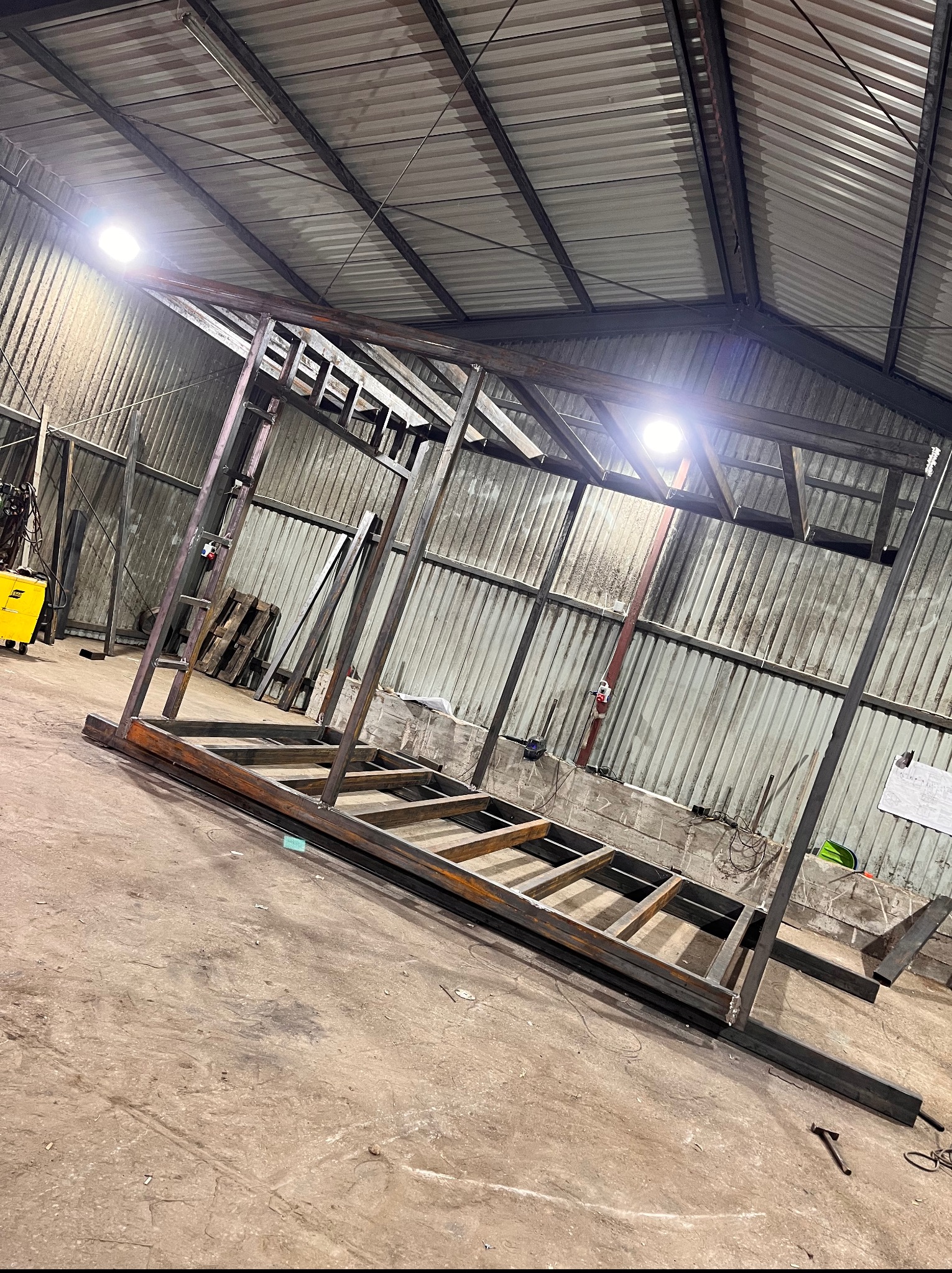 Konstrukcja stalowa hali w trakcie budowy, widoczna spawarka i fragment rysunku technicznego, surowy stan, oświetlenie przemysłowe.