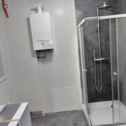 Armbud - Łazienka w trakcie prac wykończeniowych: kabina prysznicowa z szarą ścianą, biały piec gazowy na ścianie z białych płytek, częściowo zamontowana szafka pod umywalkę.