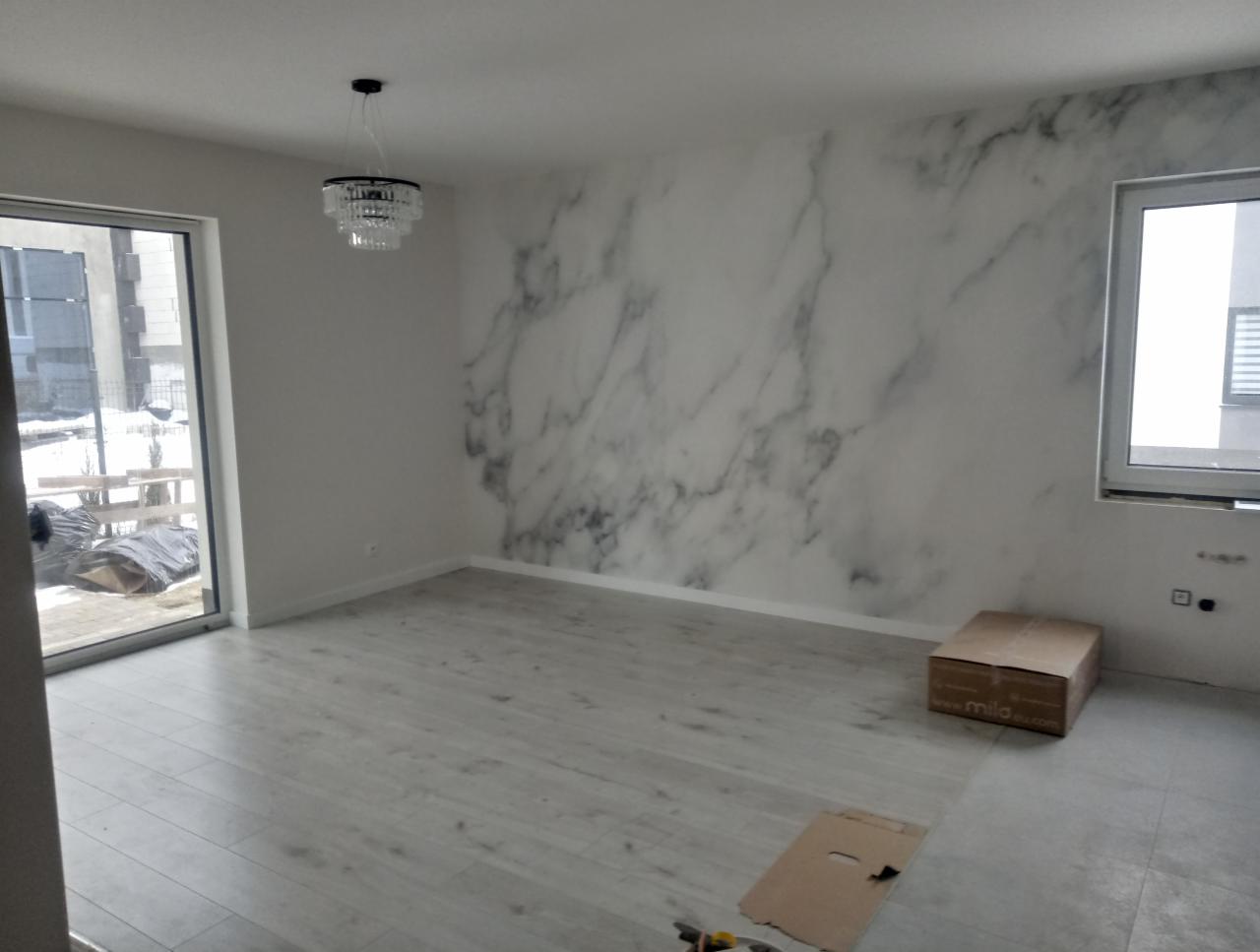 Pusta przestrzeń w trakcie wykańczania: ściana z motywem marmuru, jasna podłoga, widoczna lampa sufitowa i karton na podłodze.
