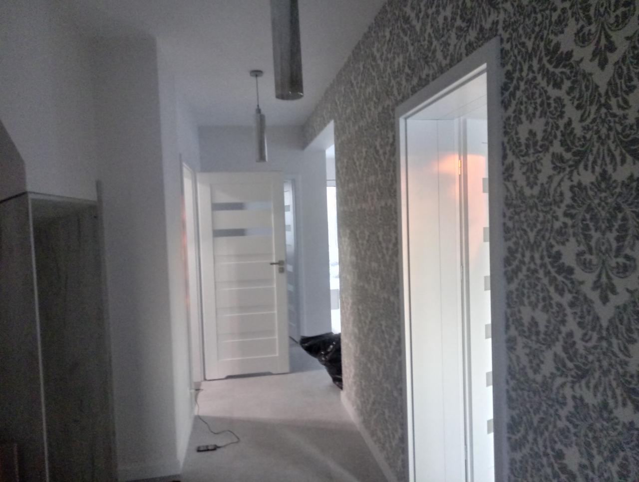 Korytarz po remoncie z szarą tapetą w ornamenty, białe drzwi z przeszkleniami i minimalistyczne, wiszące lampy.