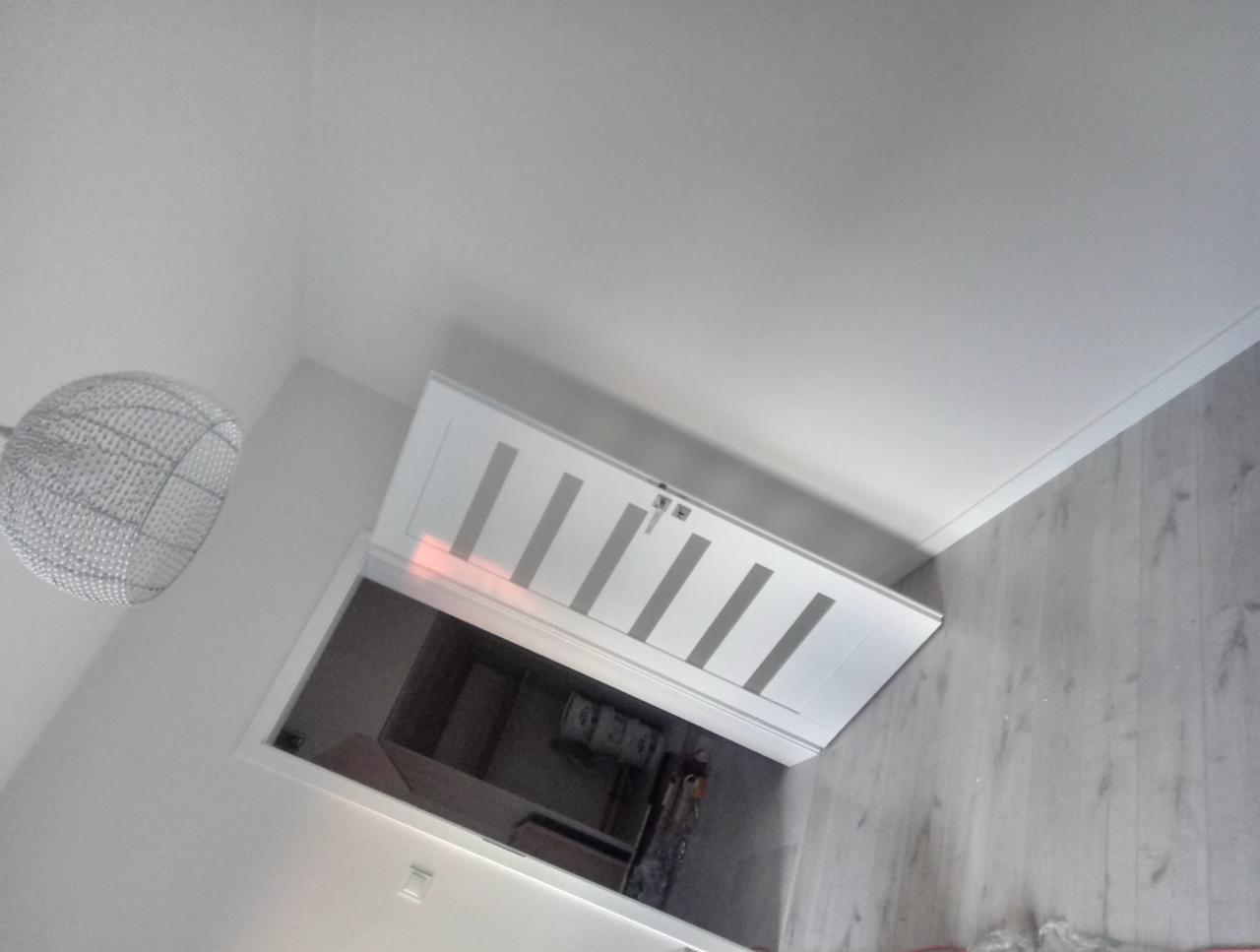 Otwarta, biała, nowoczesna klapa strychowa z pionowymi przeszkleniami, widok z dołu, lampa wisząca z metalowej siatki, szare panele podłogowe.