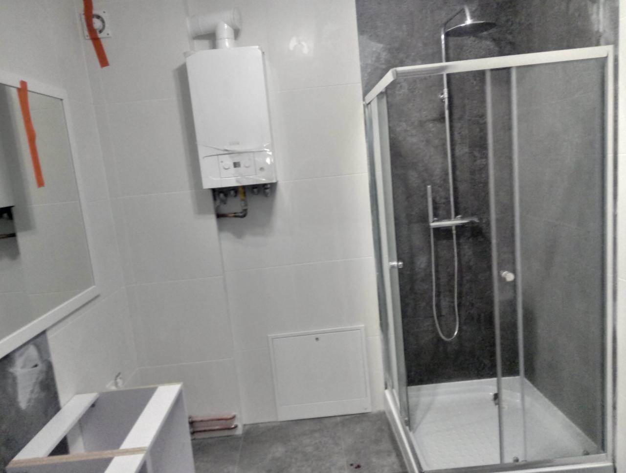 Łazienka w trakcie prac wykończeniowych: kabina prysznicowa z szarą ścianą, biały piec gazowy na ścianie z białych płytek, częściowo zamontowana szafka pod umywalkę.