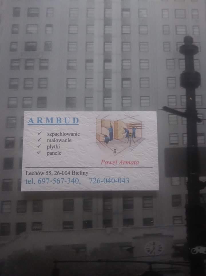 Wizytówka firmy ARMBUD z ofertą szpachlowania, malowania, układania płytek i paneli, na tle rozmazanego budynku.