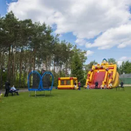 Rozległy plac zabaw na trawie z dmuchanym zamkiem ze zjeżdżalnią, tunelem z motywem motyla, trampoliną z podwójnym koszem i rodziną siedzącą na ławce w tle drzew.