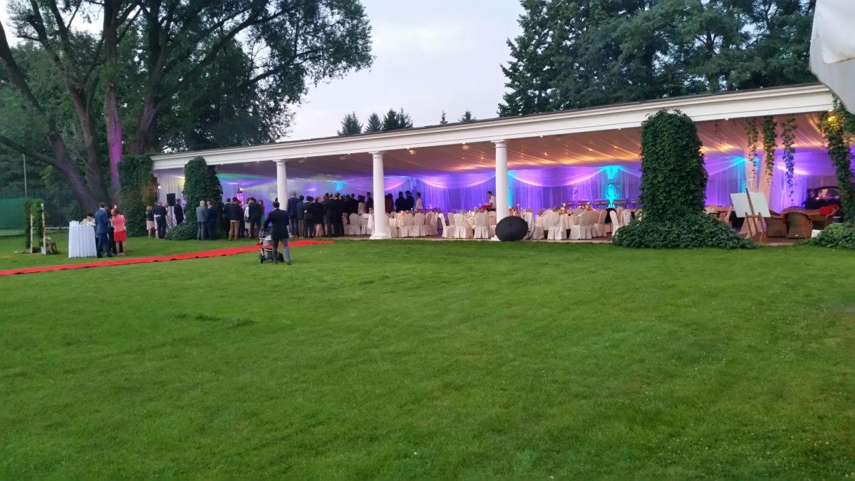 Uroczystość plenerowa z czerwonym dywanem, białym namiotem kolumnowym oświetlonym kolorowymi światłami, stołami i krzesłami, oraz gośćmi w tle.
